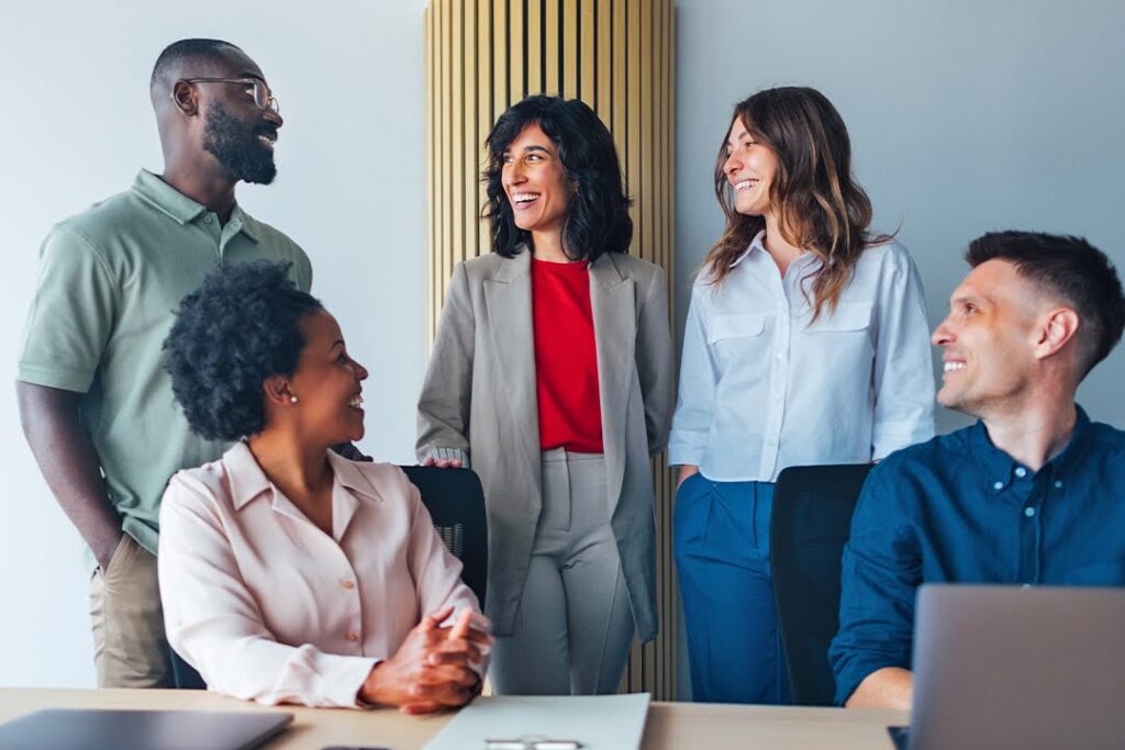 A group of five professionals smiles and talks together in an office setting, suggesting teamwork and collaboration.