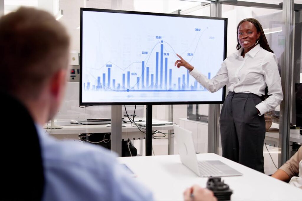 A woman presents data on a large screen with bar and line graphs to colleagues in a modern office meeting room.