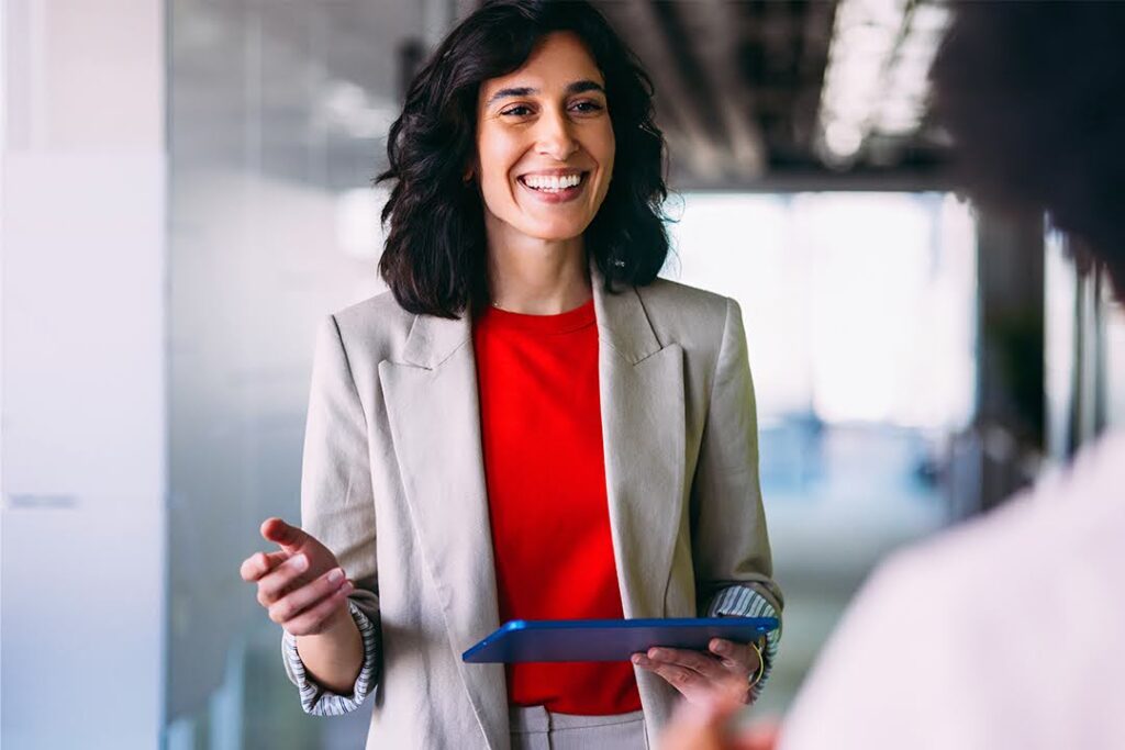 A smiling woman holds a tablet while speaking with a colleague in a bright, modern office hallway.